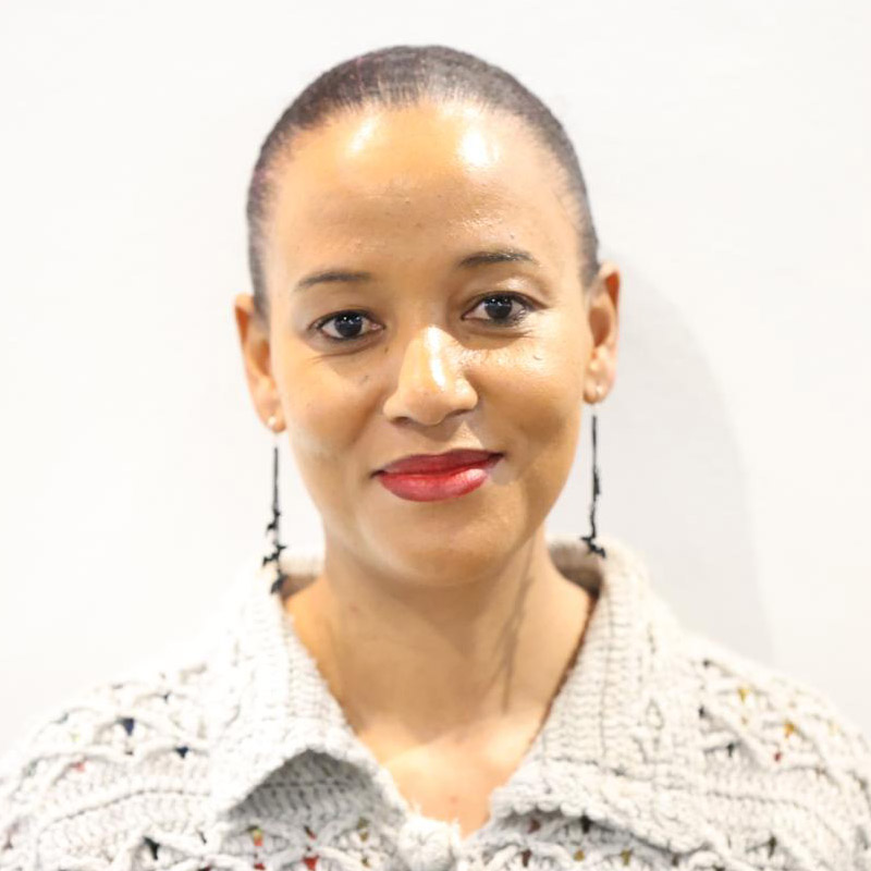 woman with dark brown hair, pulled back, and brown eyes, dangling earrings, reddish lipstick, wearing a off white knit or crocheted top. She's looking at the camera.