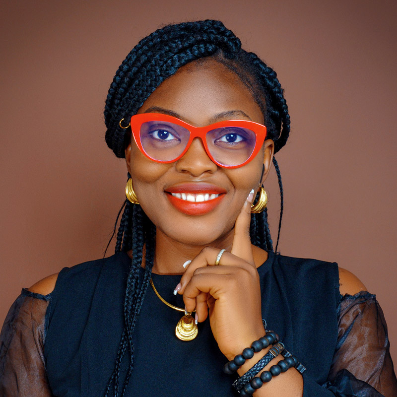 Black woman with braids, lipstick, red glasses, gold earrings and a gold necklace, black top. she's looking at the camera.